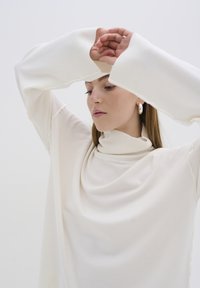 Przesadnie duży sweter w odcieniu kremowym, z wysokim dekoltem, prążkowanymi mankietami i miękką, gładką fakturą z detalami drapowanych tkanin.