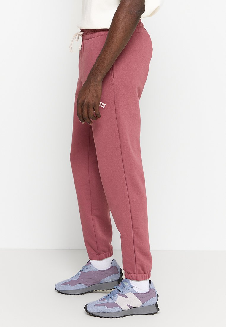 Helle rosa Jogginghose mit elastischem Bund und Kordelzug. Mit weißem Schriftzug "NICCE" am linken Oberschenkel. Getragen mit lila Turnschuhen.