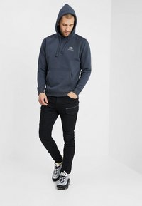 Felpa blu navy con tasca frontale e cappuccio con cordini; abbinata a pantaloni neri slim-fit con tasche con zip e sneakers grigie.