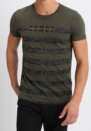 Mężczyzna w oliwkowozielonym t-shircie z krótkim rękawem, z czarnymi paskami w kamuflażu i napisem „BNM 02 Denim brand only for the bold” z przodu.