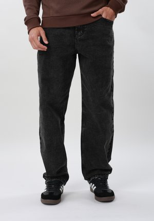 Oseba, oblečena v široke črne kavbojke, črno-bele superge in rjavo prešito jopico, z eno roko v žepu.