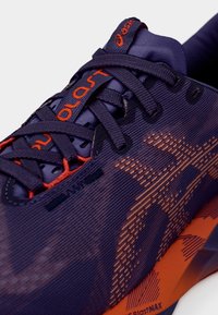 Zapato deportivo morado con detalles en naranja. Cuenta con una parte superior texturizada, patrones intrincados y cordones con marca. Diseño ligero.