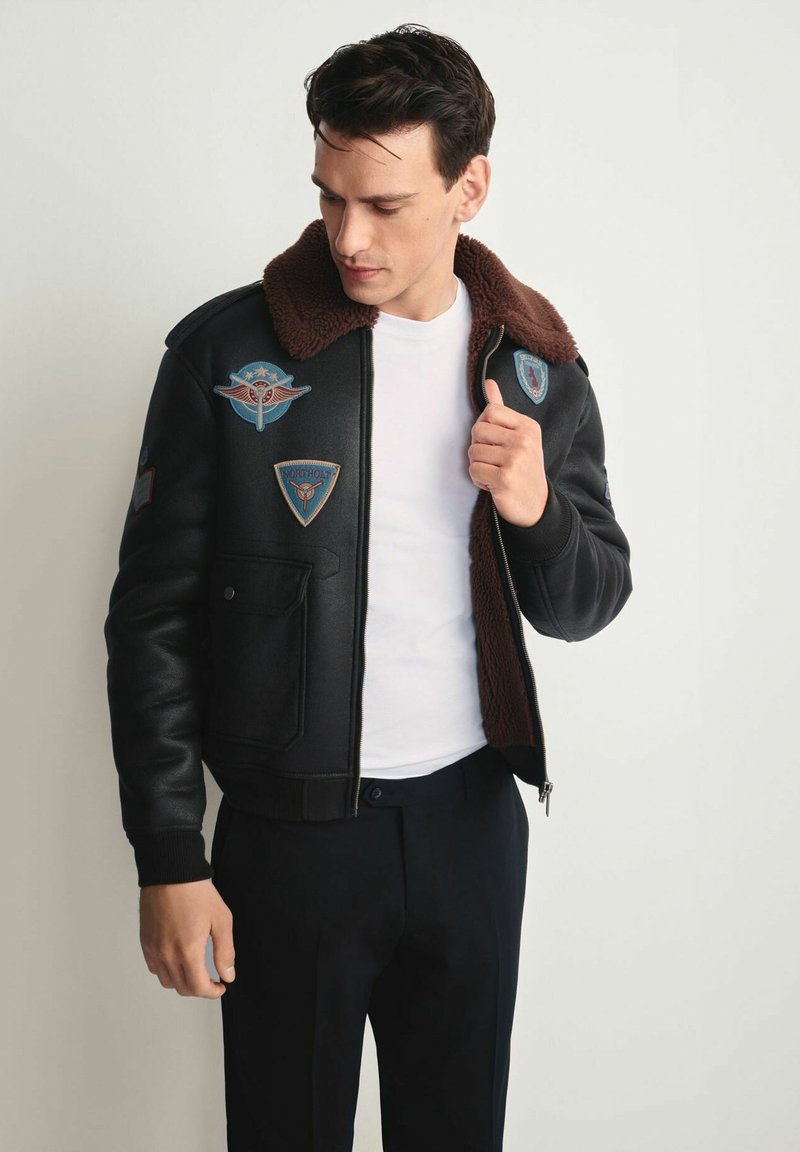 Czarna kurtka bomberka z brązowym futerkiem na kołnierzu, zamek błyskawiczny na przodzie oraz licznymi haftowanymi aplikacjami na przodzie i rękawach.