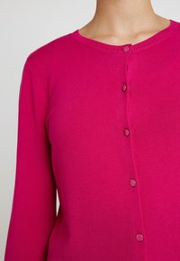Fuksiansävyinen neulejakku pyöreällä kaula-aukolla, pitkät hihat ja kuusi samanväristä nappia edessä. Sileä pinta ja istuva malli.