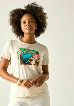 Kobieta z kręconymi włosami w beżowym t-shircie z kolorowym nadrukiem wybrzeża Capri, stojąca na jednolitym jasnym tle.