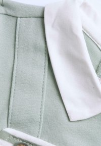 Groene stof met een gladde textuur, voorzien van een witte kraag, knoopdetails en contrasterende stiksels. Zacht en getailleerd ontwerp.