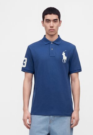 Jeune homme portant un polo bleu avec un logo blanc de joueur de polo sur la poitrine et le numéro 3 sur la manche droite.