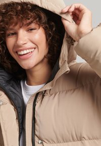 Pufferjakke i beige med sort indvendig for, udstyret med hætte, lynlås og glat, quiltet struktur. Modellen smiler med håret ned.