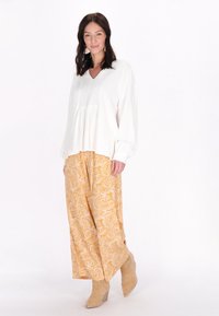 Witte blouse met lange mouwen en V-hals; gele broek met paisleymotief en wijde pijpen; lichtbeige enkellaarsjes; effen achtergrond.