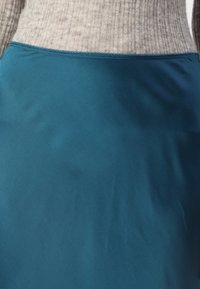 Gros plan sur une personne portant un pull côtelé beige rentré dans une jupe lisse teal, mettant en évidence les textures du tissu et les détails de couture.
