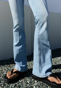 Blugi evazați de culoare albastru deschis din denim, cu cusături vizibile; asortați cu flip-flops negre. Solul este acoperit cu pietricele mici de culoare gri.
