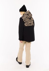 Schwarze Jacke mit Leopardenmuster auf dem Rücken, kombiniert mit beigen Hosen und schwarzen Stiefeln. Modell trägt eine schwarze Mütze und hat eine entspannte Haltung.
