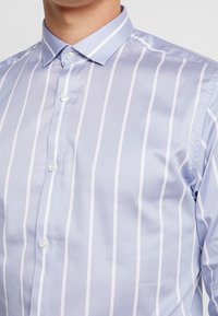 Muž v světle modré košili s bílými svislými pruhy a bílými knoflíky, zobrazený od krku do poloviny trupu.