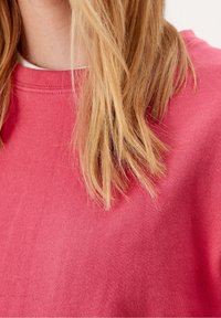 Blond vlasy odpočívají na růžovém svetru s kulatým výstřihem, odhalují límec a horní část hrudi.