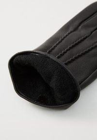Mănușă din piele neagră cu un exterior neted, căptușeală interioară texturată și detalii de cusătură discrete pe partea din spate. Design cu manșetă deschisă.