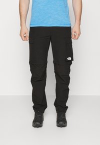 Svarta cargobyxor med flera fickor, tillverkade av lätt material, med en logotyp på högra låret. Kombinerat med svarta skor.