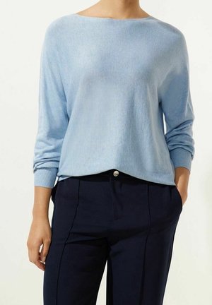 Femme portant un pull à manches longues bleu clair rentré dans un pantalon taille haute bleu marine, debout avec une main dans la poche.