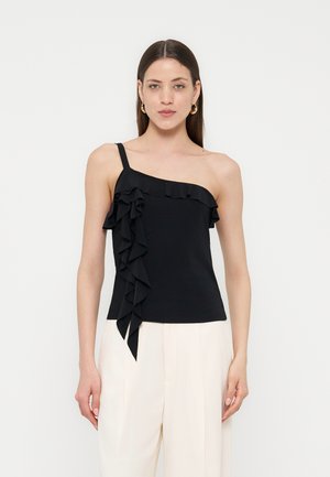 Žena u crnom topu na jedno rame s volanima i bijelim hlačama visokog struka, stoji ispred jednobojne bijele pozadine.