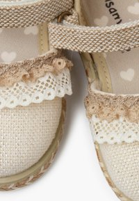 Sandalias beige con tela texturizada, adornos de encaje y correas trenzadas. Suela de goma con un detalle trenzado. Forro interior con un diseño de corazones.