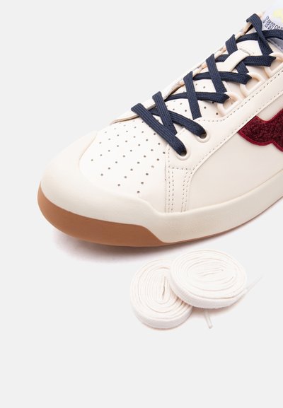 Κοντινή λήψη ενός κρεμ αθλητικού παπουτσιού με ναυτικό μπλε κορδόνια, διάτρητη μύτη, κόκκινη λεπτομέρεια στο πλάι και δύο λευκά περιτυλιγμένα ανταλλακτικά κορδόνια σε λευκό φόντο.