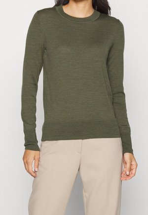 Person iført en olivengrøn strikket trøje med lange ærmer og lyse beige bukser, stående mod en ensfarvet lys baggrund.