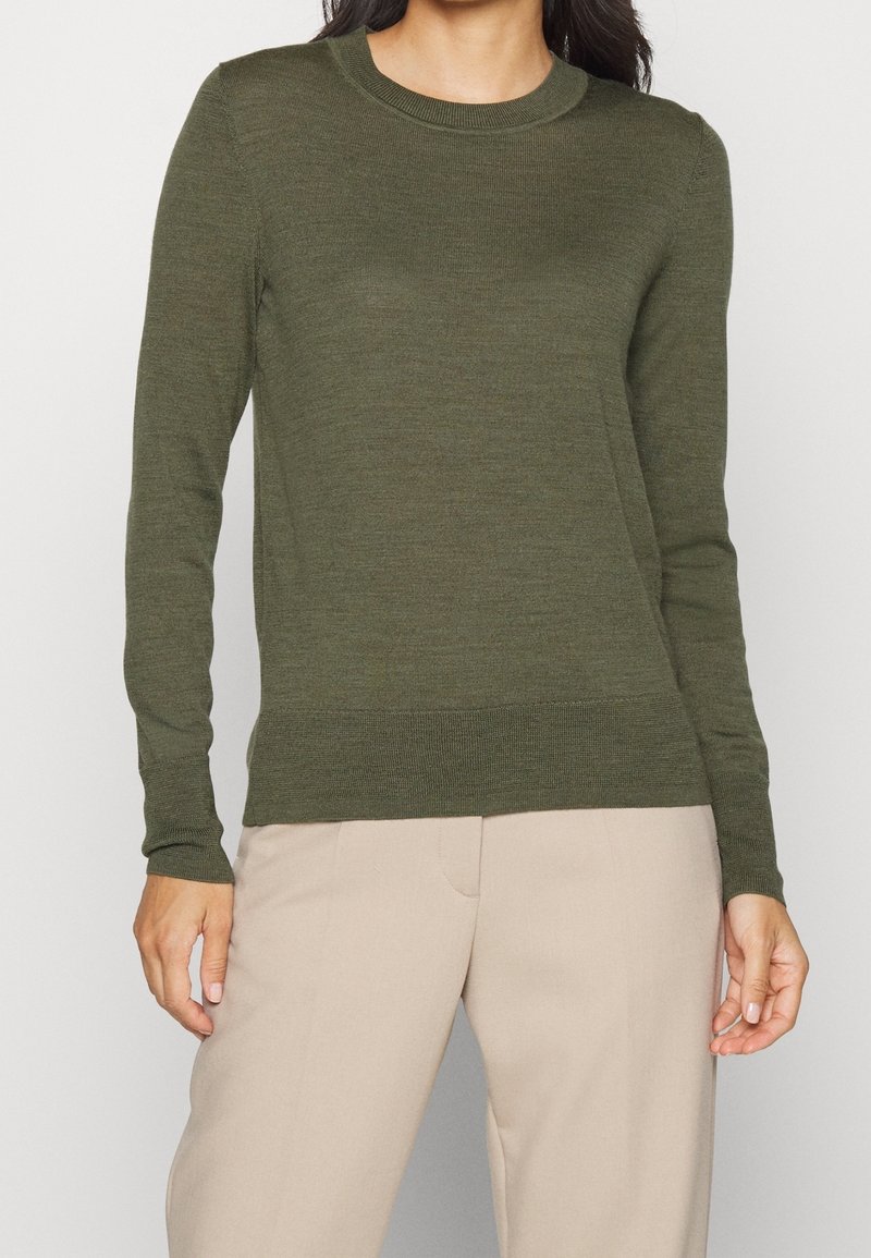 Personne portant un pull en maille vert olive à manches longues et un pantalon beige clair, debout devant un fond clair uni.
