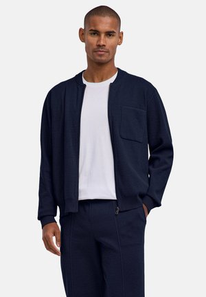 Homme portant une veste zippée bleu marine et un pantalon assorti sur un t-shirt blanc, debout avec une main dans la poche devant un fond uni.