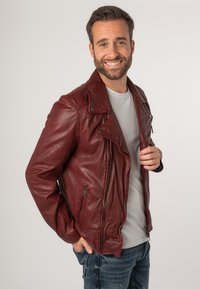 Giacca da biker in pelle bordeaux con tasche con zip, colletto a scello e finitura testurizzata, indossata sopra una t-shirt grigia chiara e jeans blu.