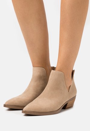 Botines de ante beige con punta afilada, laterales recortados y un tacón bajo apilado, que presentan una textura suave y detalles de costura sutiles.