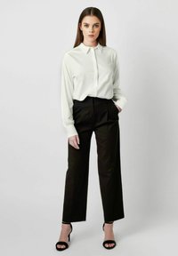Weißes Hemd mit Kragen, kombiniert mit hochgeschnittenen schwarzen Hosen. Das Modell steht mit einer Hand in der Tasche. Schwarze Riemchen-Heels.