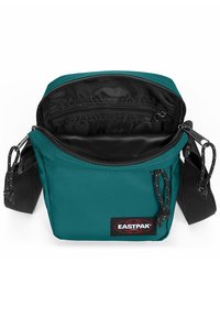 Teal Eastpak crossbody-väska med svart interiör, dragkedjeslutning, justerbar rem och logotypmärke fram. Kompakt rektangulär form.