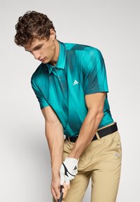 Ung man i en turkos mönstrad pikétröja och beige byxor som håller i en golfklubba med en vit handske, redo att svinga mot en enhetlig bakgrund.