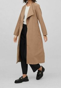 Lång brun trenchcoat utan krage, med sidfickor och rak skärning, kombinerad med svarta loafers och lösa svarta byxor.