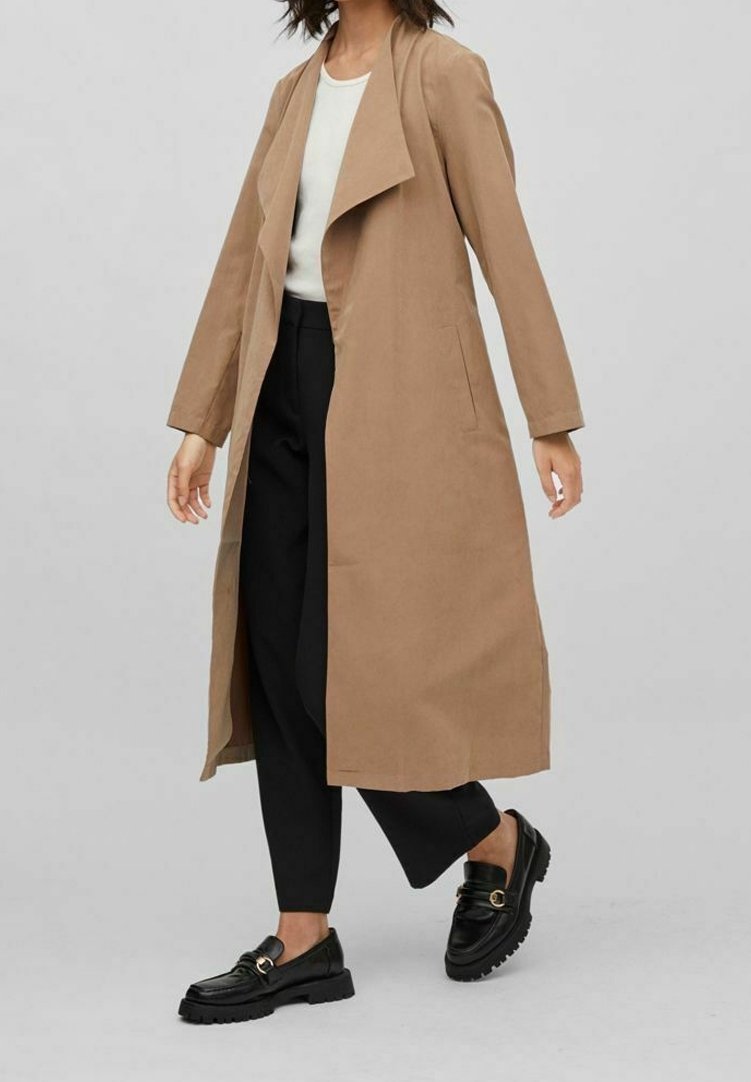 Lång brun trenchcoat utan krage, med sidfickor och rak skärning, kombinerad med svarta loafers och lösa svarta byxor.