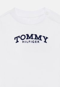 Biela bavlnená tričko s okrúhlym výstrihom a zapínaním na patentky, s námořnícky modrým vyšívaným logom "TOMMY HILFIGER" na hrudi.