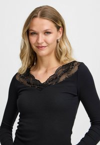 Fekete, hosszú ujjú top V-kivágással, virágos csipke díszítéssel a nyakrész körül és bordázott textúrával az egész anyagon.