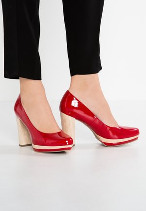 Füße in glänzenden roten Lackleder-Pumps mit hellen Holzblockabsätzen und passenden Plateaus, kombiniert mit schwarzen kurzen Hosen.
