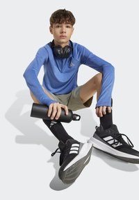 Modrá dlhá športová mikina, svetlé šortky, čierne športové topánky s bielymi pruhmi, čierne slúchadlá a čierna fľaša na vodu.