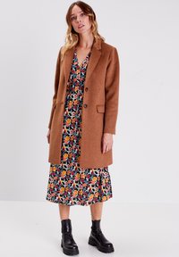 Brauner Wollmantel mit Reverskragen und zwei Knöpfen, über einem Blumendruckkleid mit leuchtenden Farben auf schwarzem Hintergrund getragen.