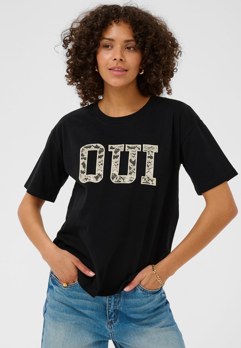 Femme aux cheveux bouclés portant un t-shirt noir avec le texte "OUI" en motif de dentelle et un jean bleu, debout les mains dans les poches.