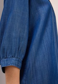 Hihna sinisestä denim-paidasta kerääntyneillä hihoilla, joka lepää juuri kyynärpään alapuolella, näyttäen kankaan tekstuuria ja ompeluyksityiskohtia.