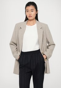 Bež blazer s teksturom i strukturiranim siluetom, s velikim reverima i dugmadi, nosi se preko bijele košulje i tamnoplavih prugastih hlača.