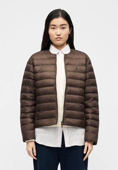 Γυναίκα που φοράει καφέ πάλλευκη ζακέτα πάνω από ένα λευκό πουλόβερ πλεχτό με σχέδιο καλώδιο και ριγέ πουκάμισο με κουμπιά, στέκεται μπροστά σε απλό φόντο.