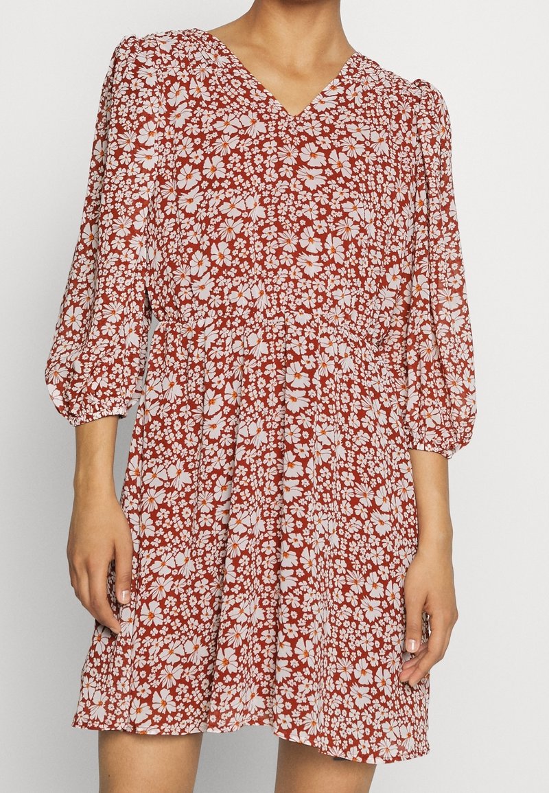 Femme portant une robe couleur rouille avec imprimé floral blanc, encolure en V et manches trois-quarts bouffantes.