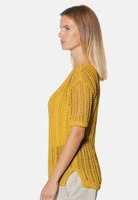 Gelber gestrickter Pullover mit kurzen Ärmeln, ausgestattet mit einer lockeren, offenen Webstruktur und Seitenschlitzen für Atmungsaktivität und Komfort.