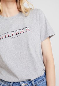 Camiseta de algodón gris con el logo "HILFIGER" en navy y rojo, con cuello redondo y mangas cortas. Corte casual, combinada con jeans azules.