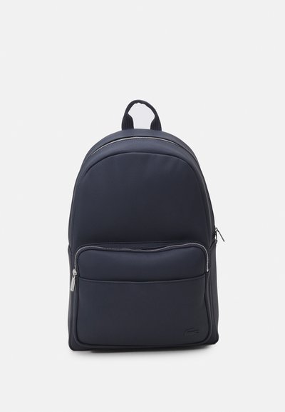 Sac à dos marine en tissu texturé avec une poche frontale lisse, des fermetures à glissière doubles et une poignée de transport supérieure. Design simple et structuré.
