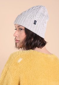 Bonnet en tricot gris clair avec un motif en côtes et une petite étiquette noire, porté avec un doux pull jaune duveteux. Le fond est neutre.
