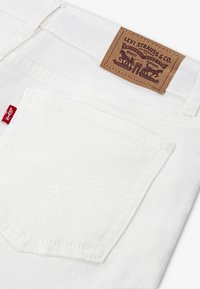 Jean en denim blanc avec une poche arrière ornée de coutures détaillées et un patch en cuir marron portant l'inscription "Levi Strauss & Co." avec un design graphique.
