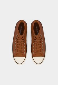 Ζευγάρι καφέ ψηλά παπούτσια Converse με κορδόνια μπροστά και λευκές λαστιχένιες μύτες, απεικονιζόμενα από πάνω σε λευκό φόντο.